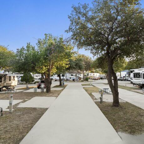 Ground-level photo of a pull-through RV site at Admiralty RV Park in San Antonio, Texas, with full hookups, picnic tables, and surrounding mature trees.
