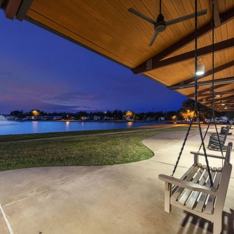 Wooden porch swing and rocking chairs on a covered patio overlooking a lit lake fountain at twilight at Lakeview RV Resort by Quality RV Resorts