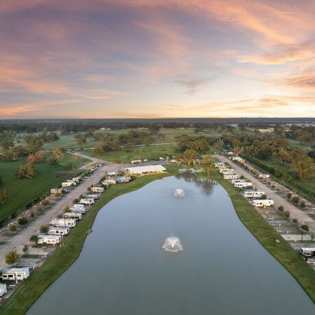 Twilight aerial view over the lake at Brazoria Lakes RV Resort in Brazoria, TX, with two fountains, clubhouse, pool, entrance, and surrounding pasture at sunset