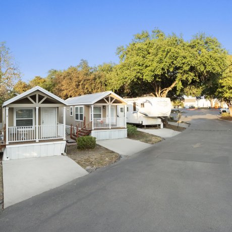 Daylight view of small park model cabins at Admiralty RV Park in San Antonio, Texas, with front porches, nearby RV sites, and tree-lined streets.