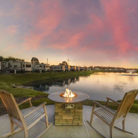 Two wooden rocking chairs by a fire pit overlooking a fountain-lit pond at sunset at Fallbrook RV Resort.