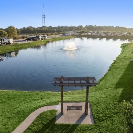 Lakeside swing under a pergola at Fallbrook RV Resort with RVs parked around a tranquil water fountain in Houston, Texas.
