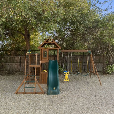 Wooden playground set with slide, swings, and climbing wall at Admiralty RV Park in San Antonio, Texas, surrounded by trees and gravel ground cover.
