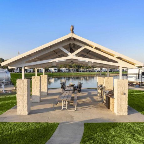 Covered picnic area and BBQ pavilion next to lake at Fallbrook RV Resort in North Houston with grills and shaded seating.