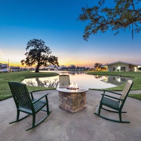 Firepit with fire and three rocking chairs at Brazoria Lakes RV Resort, with lake, sunrise, and lakefront RVs in the background in Brazoria, Texas