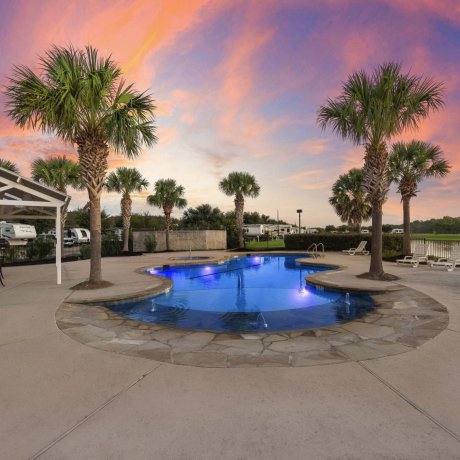 Twilight view of illuminated pool with palm trees, lounge chairs, and cabana at Westlake RV Resort in Houston TX