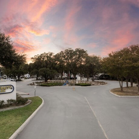 Driveway entrance of Admiralty RV Park in San Antonio, Texas, at sunset with visible signage, roundabout, and RVs beneath tall oak trees.