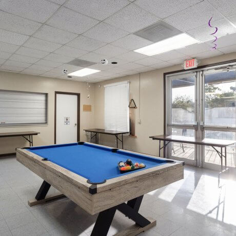 Indoor recreation room at Admiralty RV Park in San Antonio, Texas, featuring a modern blue felt pool table, cue rack, folding tables, and double-door patio access.