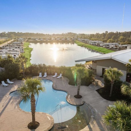 Aerial view of the swimming pool and scenic lake at Fallbrook RV Resort in Houston, TX, surrounded by RVs and palm trees on a sunny morning.