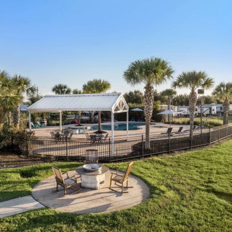 Firepit area at Eastlake RV Resort in Houston, TX with rocking chairs, pool in the background, palm trees, and professionally landscaped green lawn
