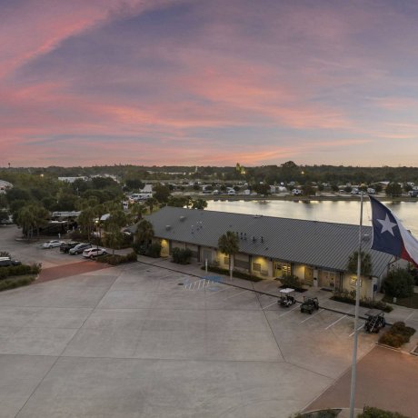 Aerial view of the main office and registration building at Lakeview RV Resort at sunset, featuring a large paved parking lot and flying American and Texas flags.