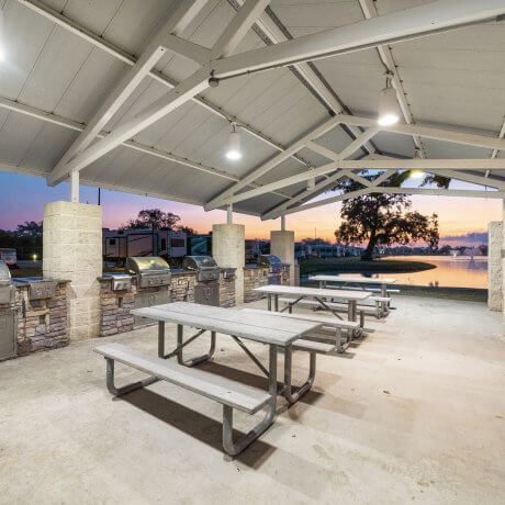BBQ area at Brazoria Lakes RV Resort with four stainless steel grills, three picnic tables, and a lake with sunrise in the background in Brazoria, Texas