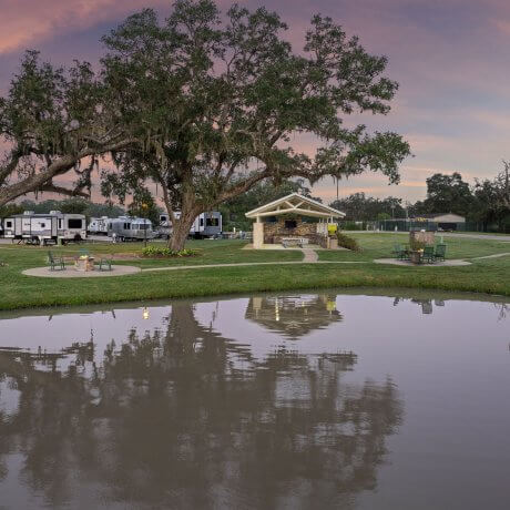 View over the lake at Brazoria Lakes RV Resort looking toward the BBQ area, oak trees, and outdoor fireplace in Brazoria, Texas
