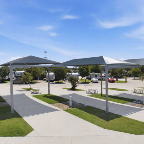 Ground-level wide view of premium covered RV camping sites featuring concrete parking pads, large shade canopies, grass medians, and utility hookups under a blue sky.