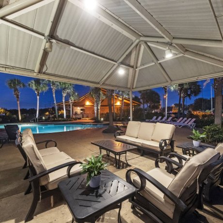 Covered lounge seating under a pavilion overlooking the illuminated resort-style pool and palm trees at Lakeview RV Resort in Houston at twilight.